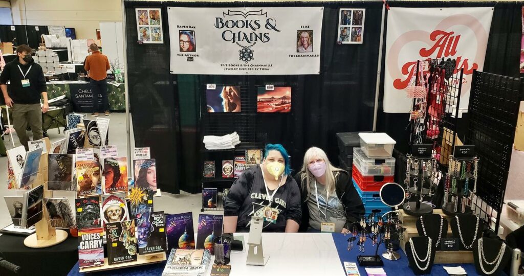 Photo of Raven Oak and Elise sitting at their Books & Chains booth at GeekGirlCon 2024. 